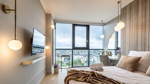 Habitación de hotel moderna con una cama grande, un televisor montado en la pared y vistas a la ciudad a través de amplios ventanales.