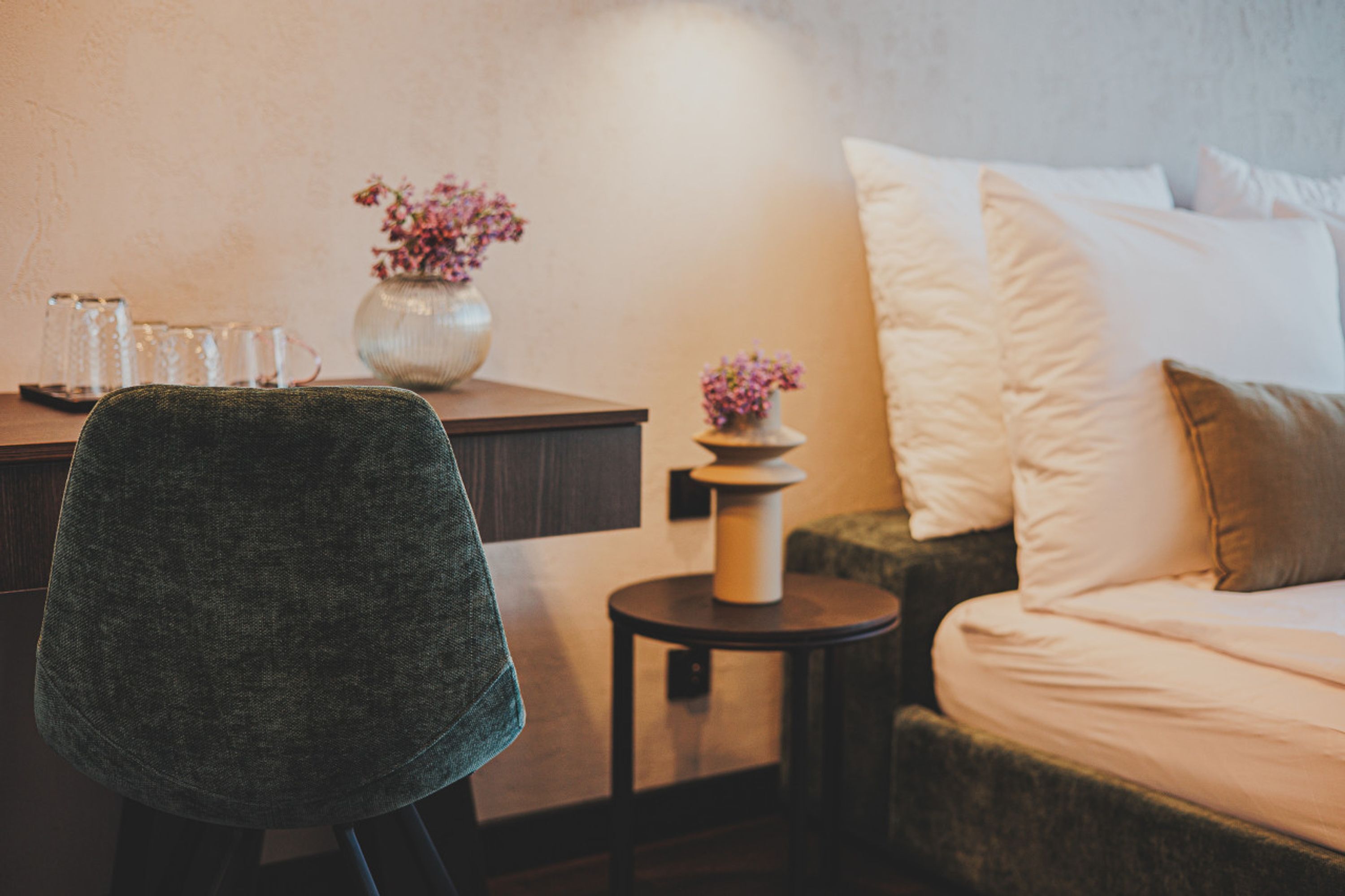 A cozy hotel room with a bed, a small round table with a vase of flowers, and a chair.