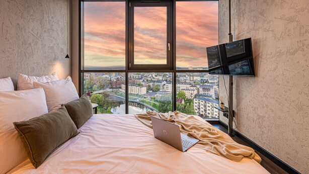 Een slaapkamer met een groot raam, een bed, een laptop en een televisie aan de muur.