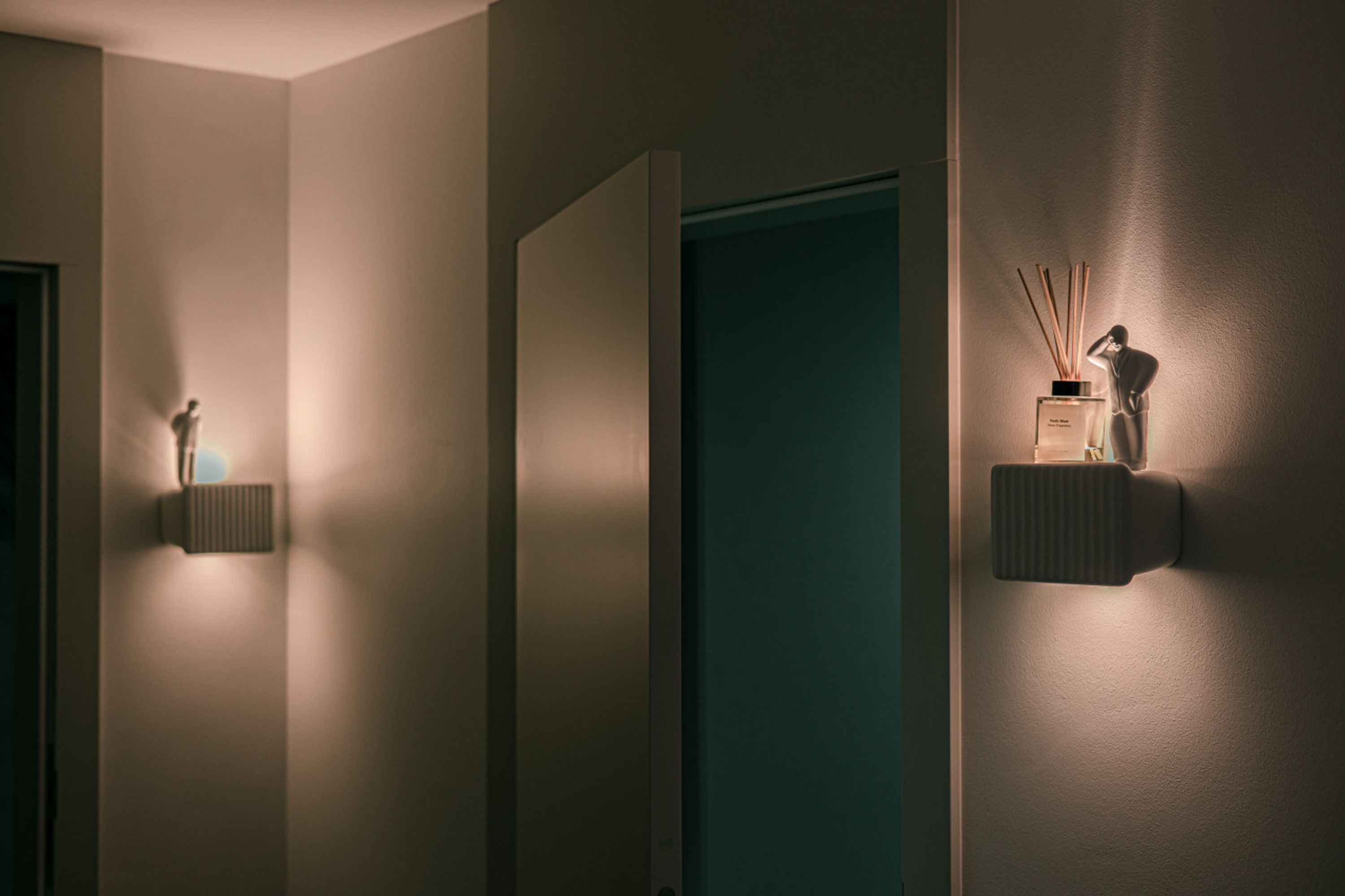 A hallway with a teal door, a wall-mounted light fixture, and a small table with a bottle and reed diffuser.