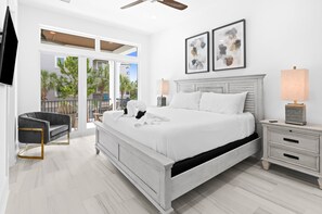 A bedroom with a large bed, a chair, a nightstand, and a window with a view of palm trees.