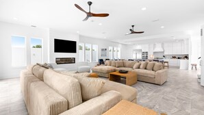 Ein großzügiges Wohnzimmer mit einer großen, beigefarbenen Eckcouch, einem Couchtisch und einem an der Wand befestigten Fernseher. Deckenventilatoren, eine Küchenzeile mit weißen Schränken und große Fenster, die für natürliches Licht sorgen, sind ebenfalls vorhanden.