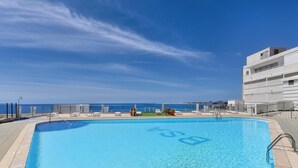 Piscina retangular com água azul cristalina, rodeada por espreguiçadeiras e vista para o mar.