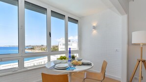 Uma área de refeições com uma mesa posta para dois, janelas amplas com vista para o mar e um abajur em um tripé.