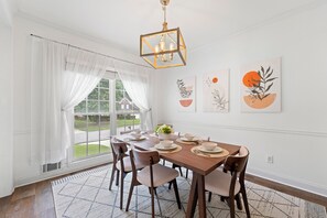 A dining room with a wooden table set for six, white curtains, and two wall art pieces. - 🌟 MODERN 5-BEDROOM🛏️, PERFECT FOR LARGE FAMILIES! SPACIOUS SUNROOM ☀️ (Lawrenceville)