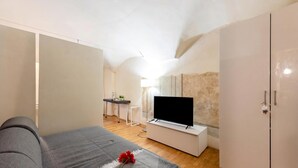 Et moderne soveværelse med en sofa, et fjernsyn på et stativ, et skab og et skrivebord med en lampe.