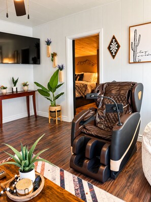 A modern living room with a massage chair, a flat-screen TV, wooden flooring, and decorative plants. - Firepit | off I-80 | Private Carport |1BR | Pets (Lyman)