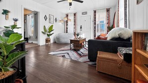 A cozy living room with a dark sofa, a wooden coffee table, and a ceiling fan. There are potted plants and a patterned rug on the wooden floor.