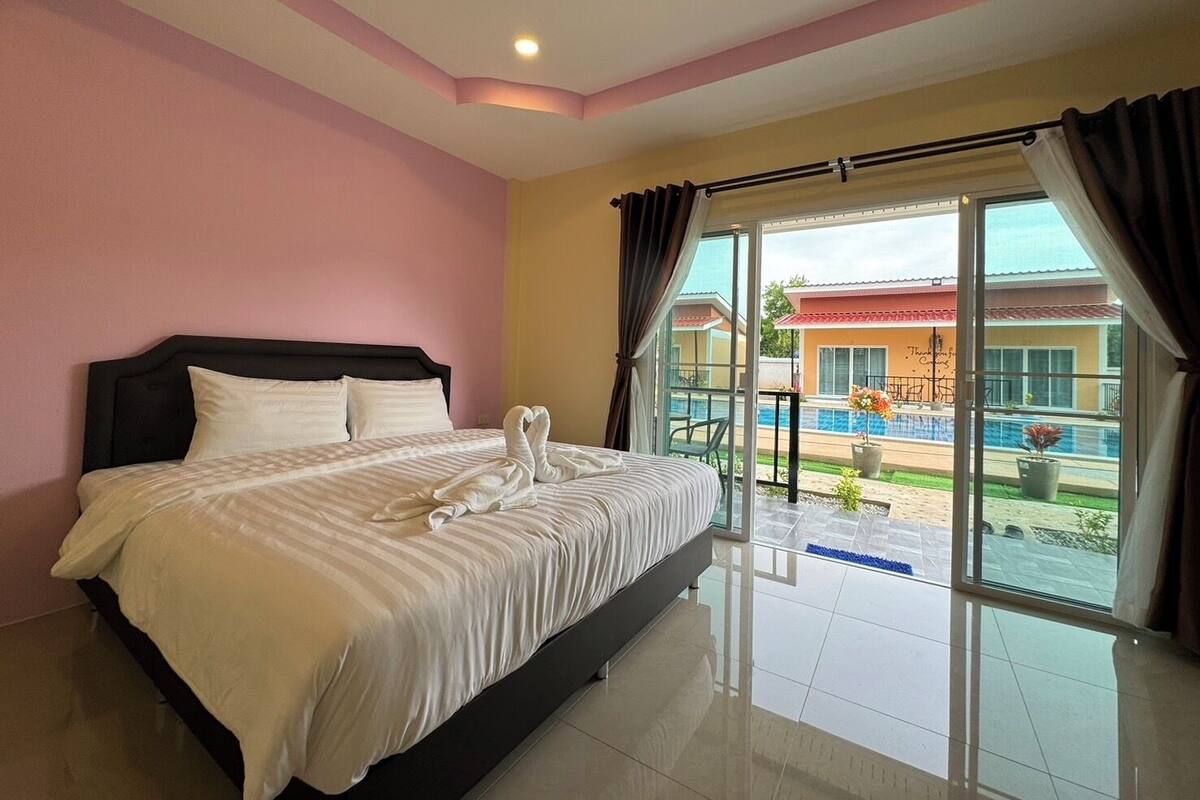 Quarto de hotel com cama, uma janela grande com cortinas e vista para uma piscina e um edifício lá fora.
