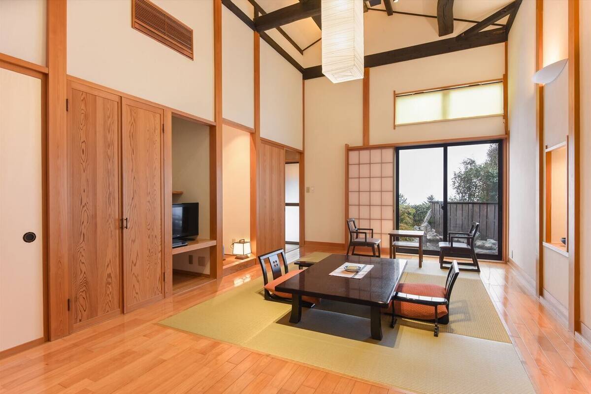 A traditional Japanese-style room with tatami flooring, a low table with cushions, sliding doors, and a large window providing a view of the outdoors.