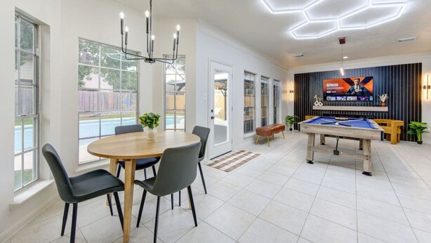 Een moderne, overdekte ruimte met een pooltafel, een eethoek met stoelen en een plafond met geometrische verlichtingsarmaturen.