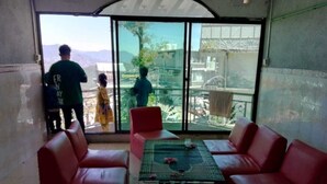 Quarto com sofás de couro vermelho, uma mesa de vidro com flores e vista para uma varanda com pessoas lá fora.