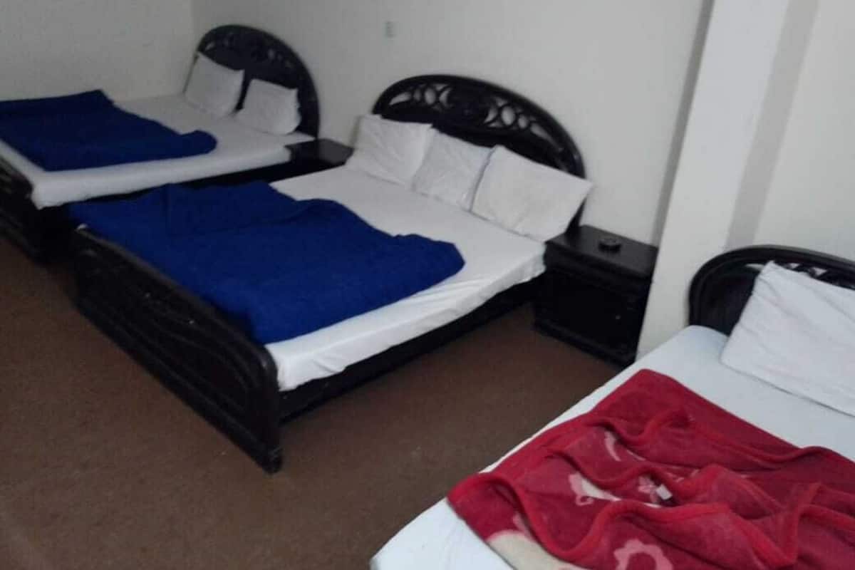 Two single beds with black frames and white bedding, a small bedside table with a lamp, and a brown carpeted floor.