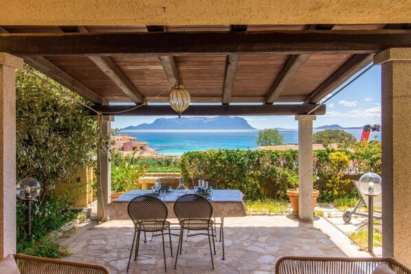 Une terrasse extérieure avec une table et des chaises, un luminaire suspendu et une vue sur la mer et les montagnes.