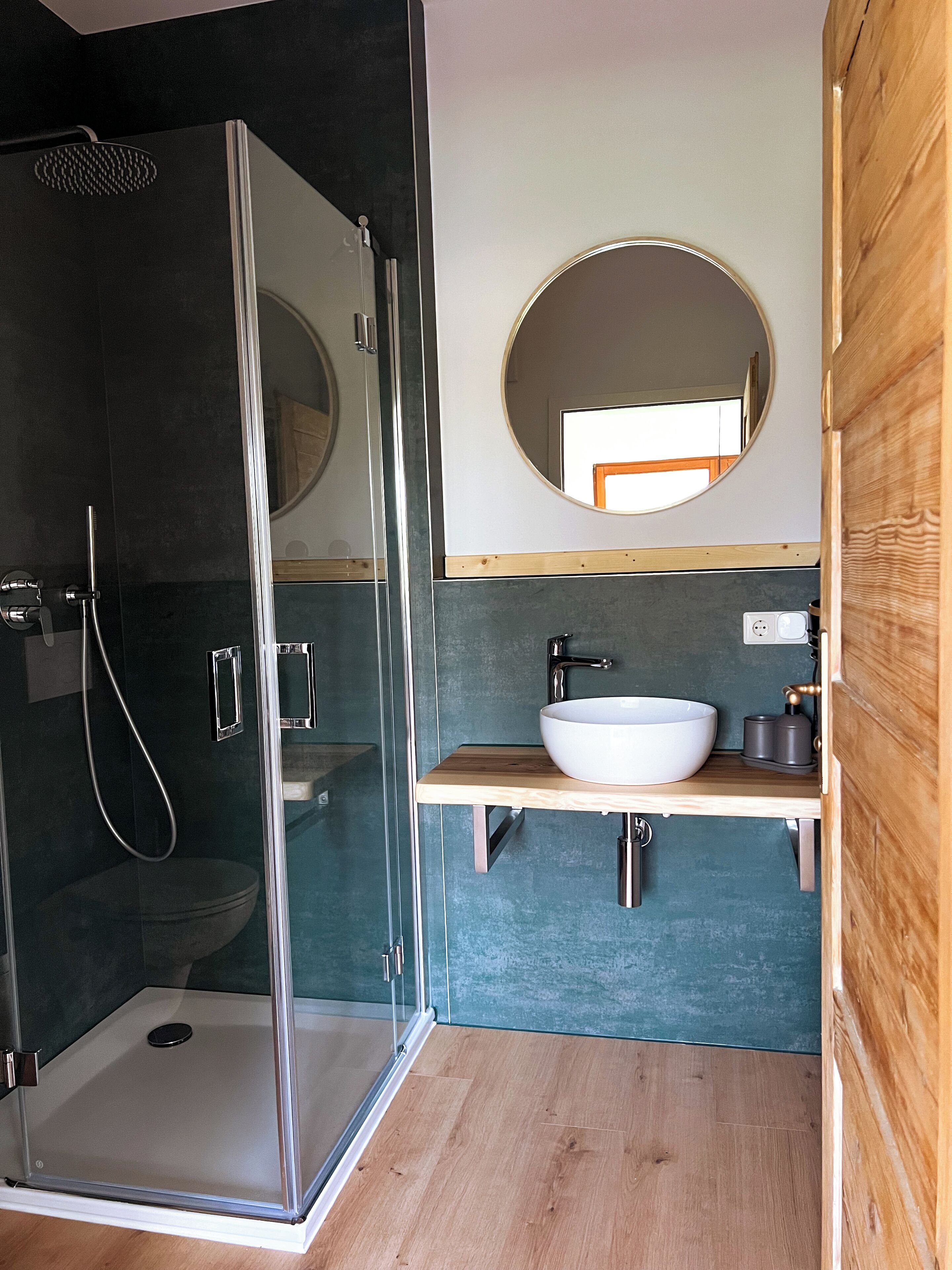 Une salle de bain moderne avec une douche à l’italienne en verre, un meuble-lavabo en bois avec un lavabo blanc et un miroir rond au-dessus du lavabo.