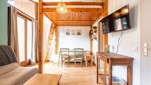 A cozy room with a wooden ceiling, a TV mounted on the wall, a dining area with chairs and a table, and a sofa with a cushion. - Nice apt for 6 people near the slopes (Morillon)