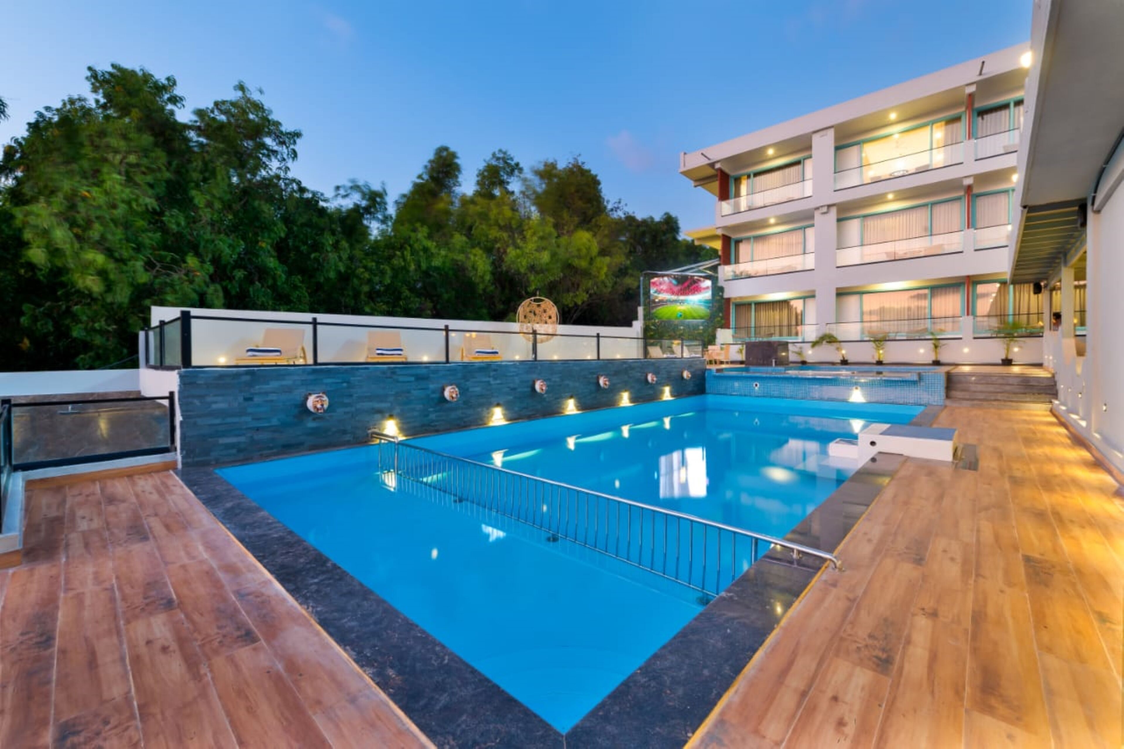 Una piscina con plataforma de madera, rodeada por un edificio moderno con varios balcones y una pantalla grande mostrando un evento deportivo.