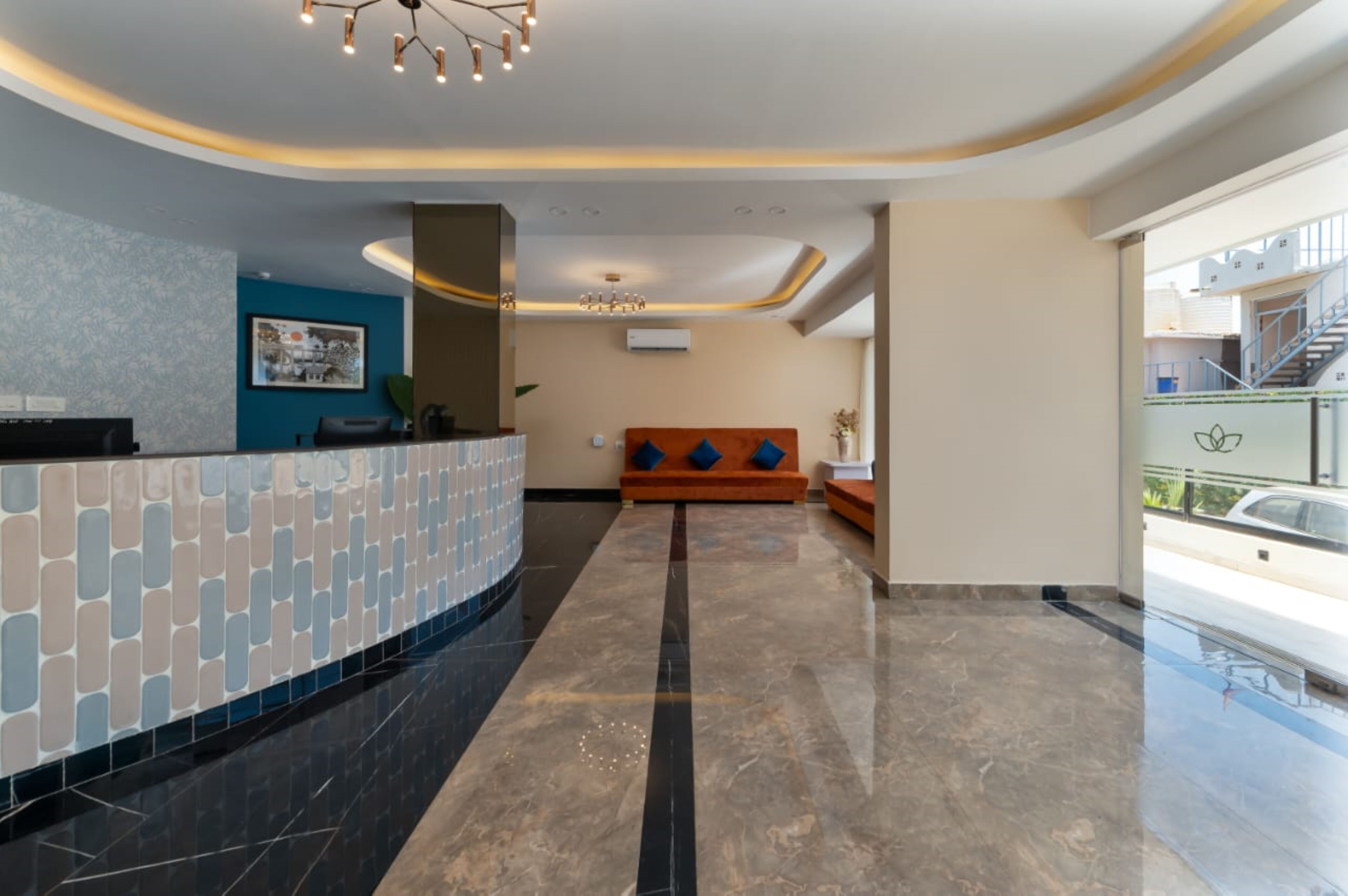 A modern hotel lobby with a curved reception desk, a blue wall with a framed picture, a seating area with orange sofas, and a glass railing overlooking the street.