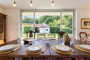 Een eettafel gedekt voor zes personen met witte borden, bestek en wijnglazen, met uitzicht op een tuin en een stenen gebouw.