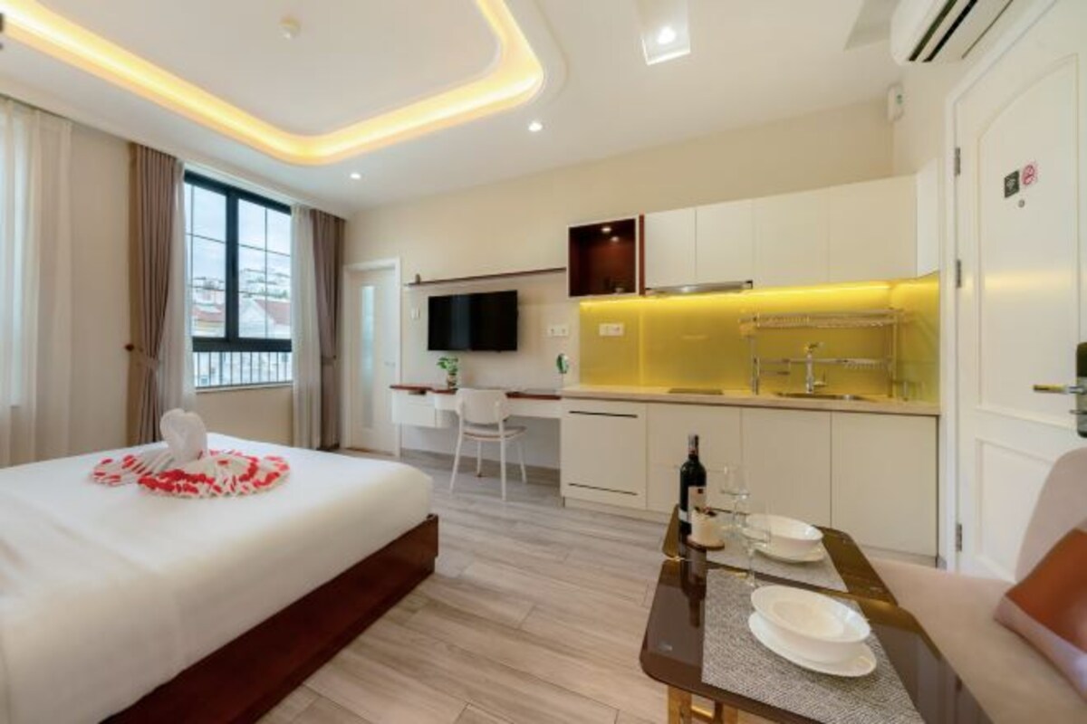 Quarto de hotel moderno com cama, mesa de jantar pequena, televisão e cozinha compacta.