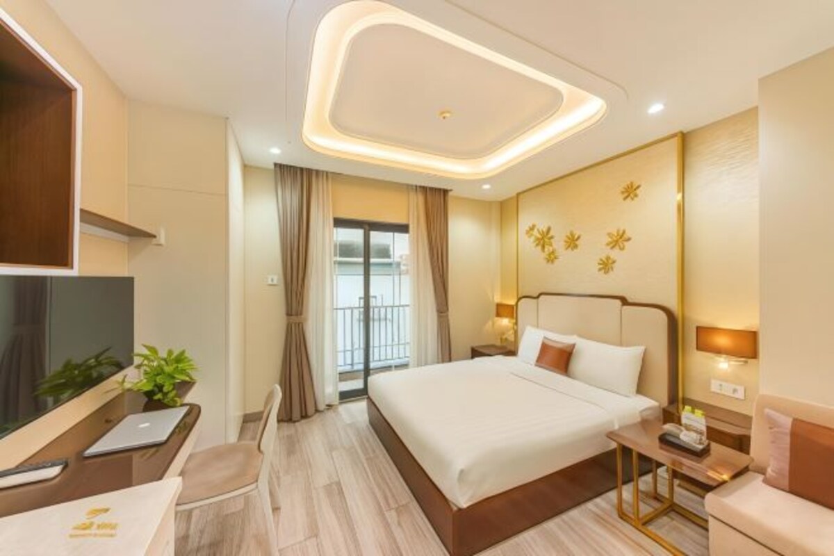 Quarto de hotel com cama, mesas de cabeceira, escrivaninha com cadeira, televisão e janela com cortinas.