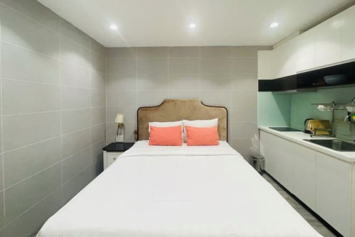 Quarto de hotel com cama, mesa de cabeceira, pequena mesa e cozinha compacta.