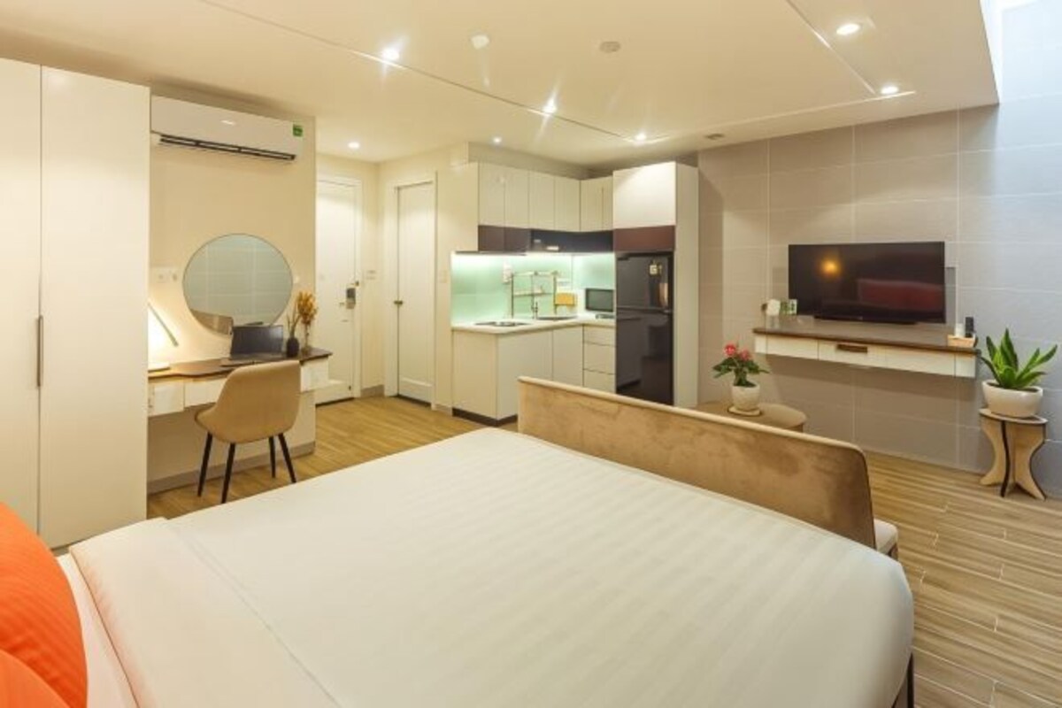 Quarto de hotel moderno com cama grande, mesa de trabalho com cadeira, área de cozinha e televisão fixada na parede.
