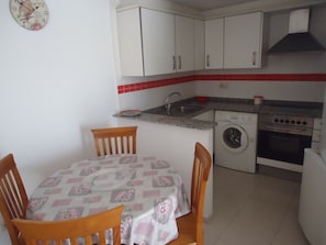 Une cuisine compacte avec des meubles blancs, un lave-linge, un four et une table à manger recouverte d’une nappe à motifs floraux.