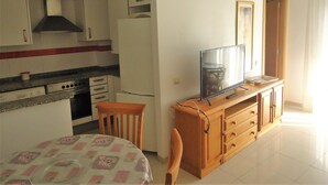 A compact kitchen with white cabinets, a stove, and a refrigerator. There's a dining table with a floral tablecloth and a chair. A television is on a wooden cabinet. - Orlando LEK (Benicarló)