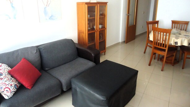 A living room with a sofa, ottoman, wooden cabinet, dining table with chairs, and a door leading to another room. - Orlando LEK (Benicarló)