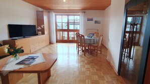 A spacious living area with a wooden ceiling, a dining table with chairs, a TV on a cabinet, and a large window with curtains.