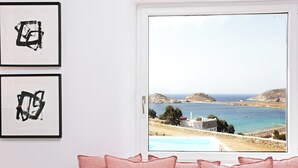 A room with a view of a coastal landscape through a window, framed artwork on the wall, and a pink cushion on the sofa. - Waterfront Villa Angelika Pink Pelikan (Mykonos)