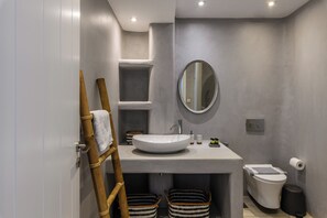 Ein modernes Badezimmer mit einem großen weißen Waschbecken, einem runden Spiegel, einer Toilette und einem Mülleimer. Unter dem Waschbecken befinden sich zwei Körbe und ein Handtuch an einer Holzleiter.