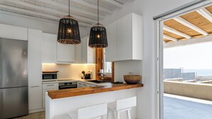 A modern kitchen with white cabinets, a wooden countertop, two hanging wicker pendant lights, and a view of the sea through a sliding glass door. - Waterfront Villa Angelika Pink Pelikan (Mykonos)