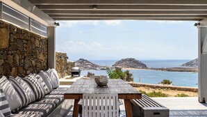 A covered outdoor seating area with a stone wall, a wooden table, and a view of the sea and hills. - Waterfront Villa Angelika Pink Pelikan (Mykonos)