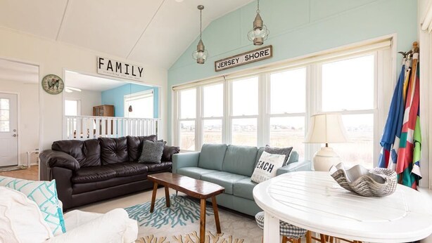 Un salon avec un canapé en cuir marron, un canapé turquoise, une table basse blanche et une table ronde blanche avec des chaises. Deux panneaux sont accrochés au mur, sur lesquels on peut lire les mots « FAMILLE » et « JERSEY SHORE ».