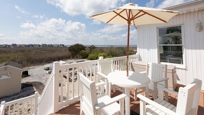 Une terrasse extérieure blanche, avec une table ronde et des chaises, un grand parasol, et une vue sur la plage et l’océan.