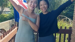Two people on a wooden deck with a flag, a bench, and greenery in the background.