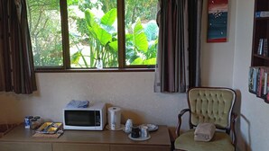 A room with a wooden desk, a microwave, a kettle, and a chair. There is a window with curtains, and a bookshelf with books.