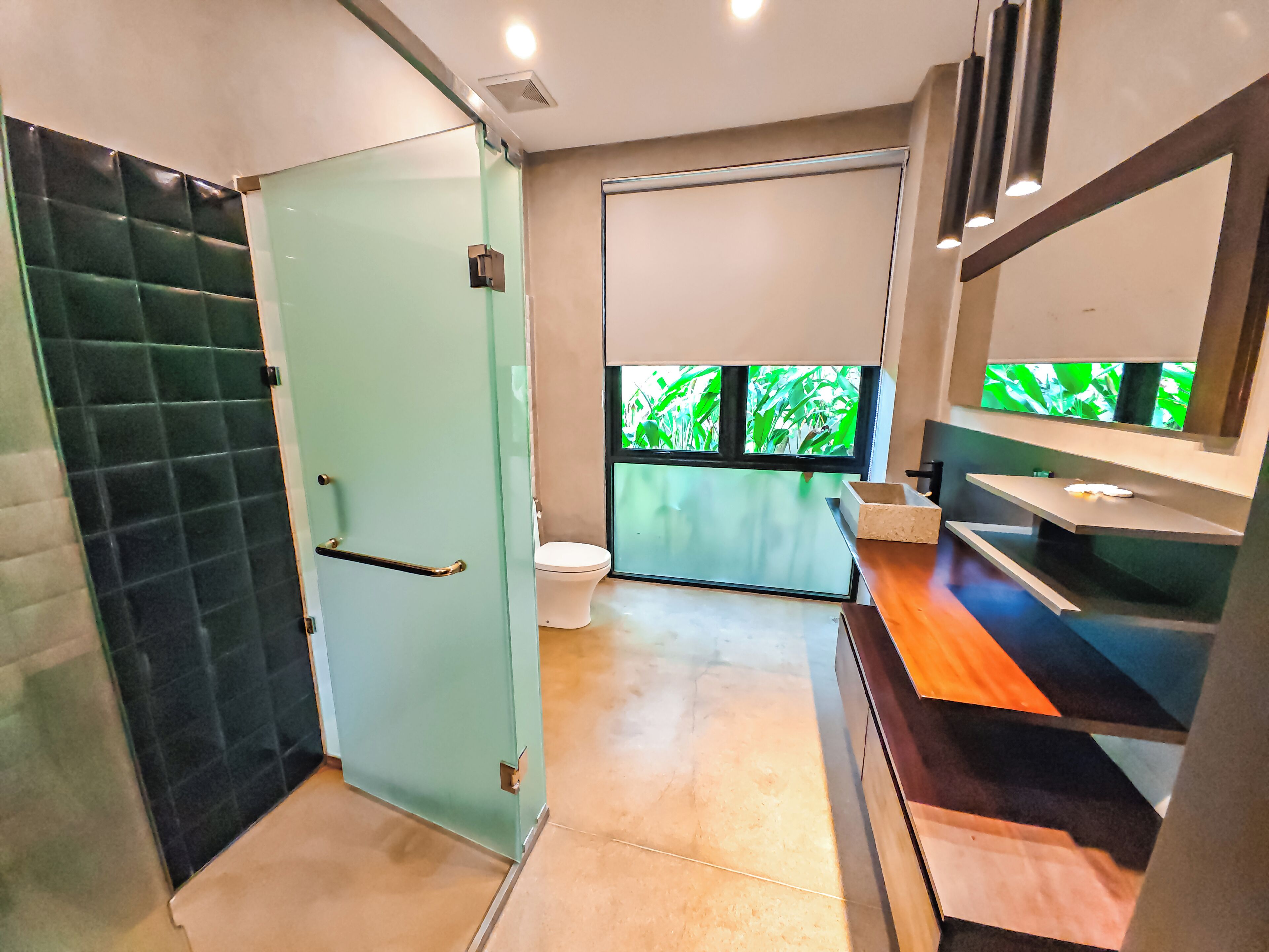 Et moderne badeværelse med glasdør, toilet, håndvask med spejl og et vindue med udsigt over grønne områder.