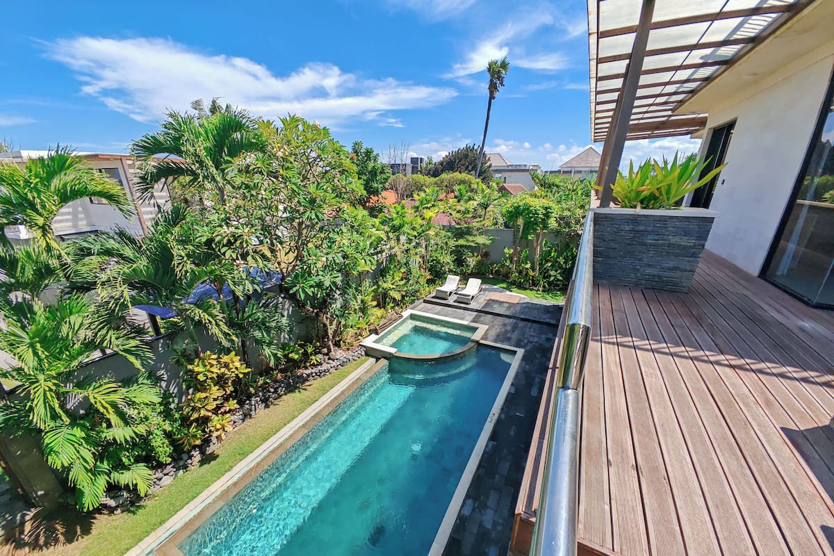 A rectangular swimming pool with a wooden deck on one side, surrounded by lush greenery and a clear blue sky.