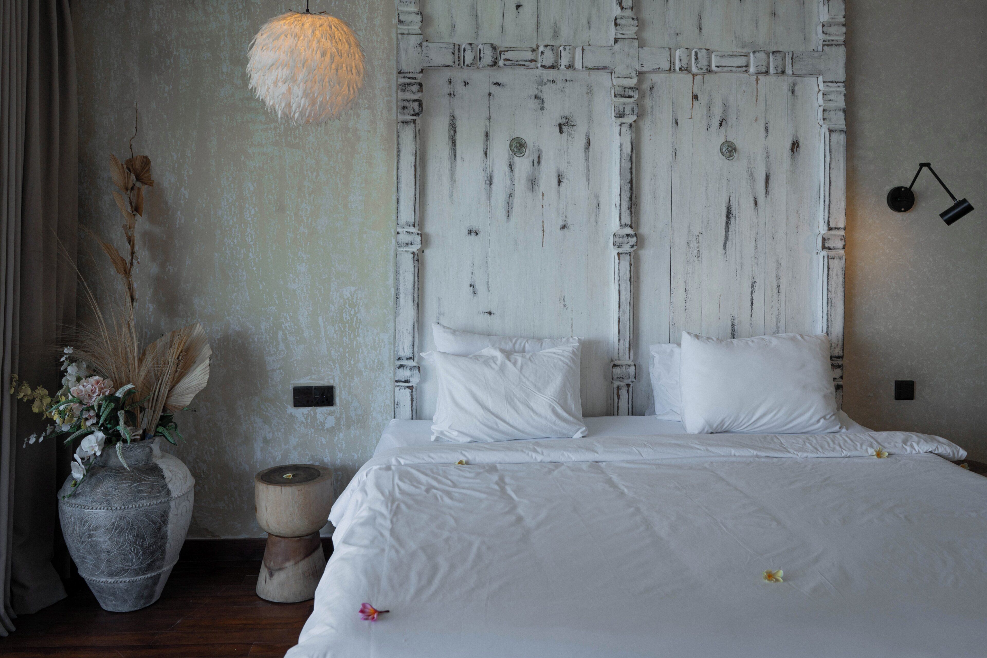 A bedroom with a large bed, white bedding, a decorative vase with dried plants, and a wall-mounted lamp.