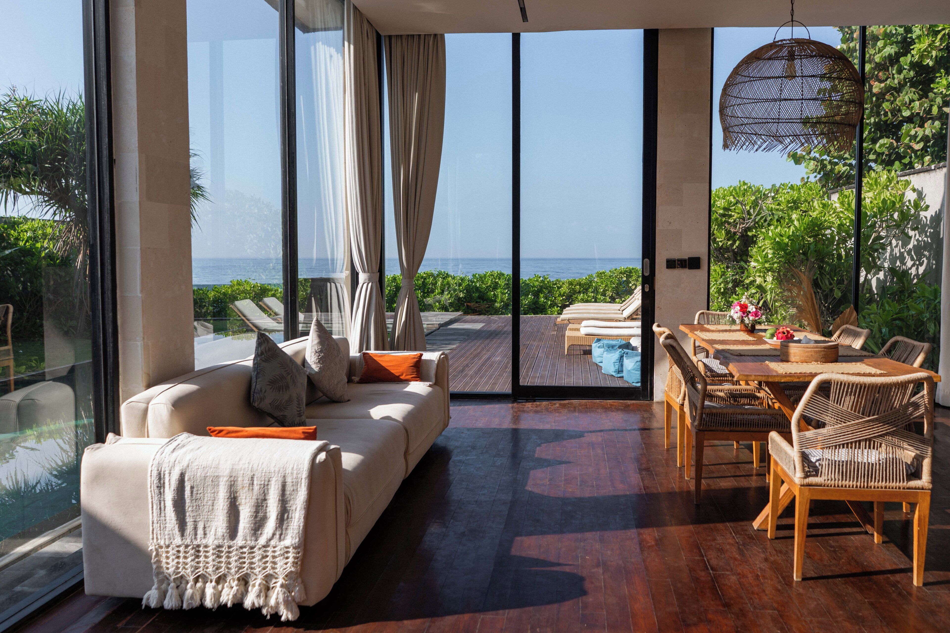 Una sala de estar moderna con un amplio ventanal que da a una zona de piscina, un sofá con cojines, una mesa de comedor con sillas y una lámpara colgante.