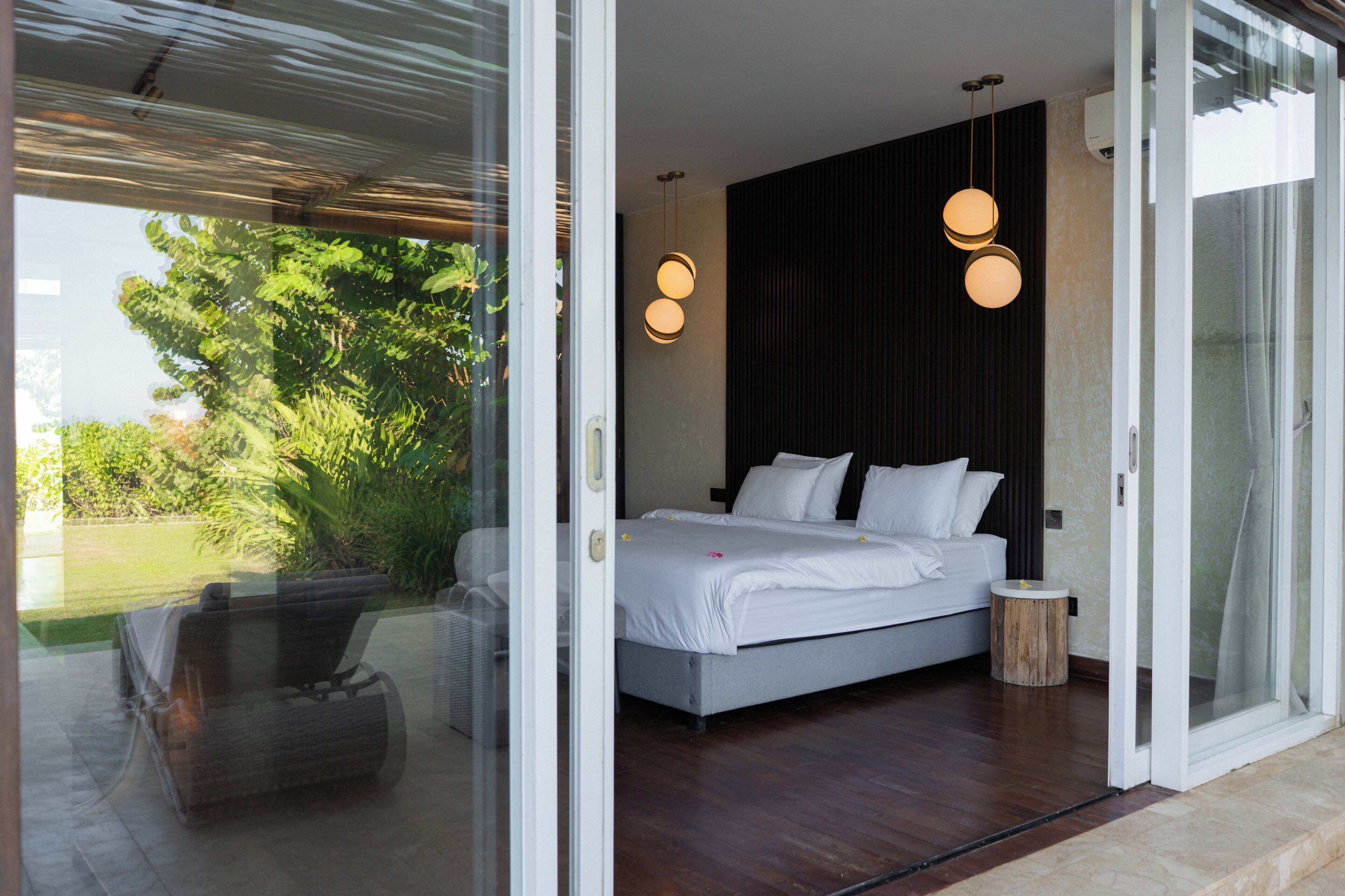 A modern bedroom with a large bed, a small round side table, and a view of greenery outside.