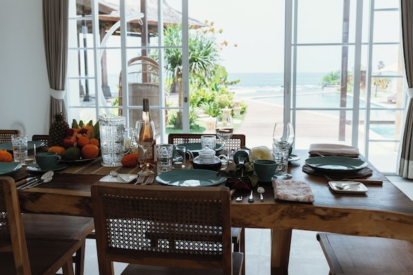Une table à manger dressée avec des assiettes, des verres et une carafe, donnant sur une piscine et offrant une vue sur l’océan.