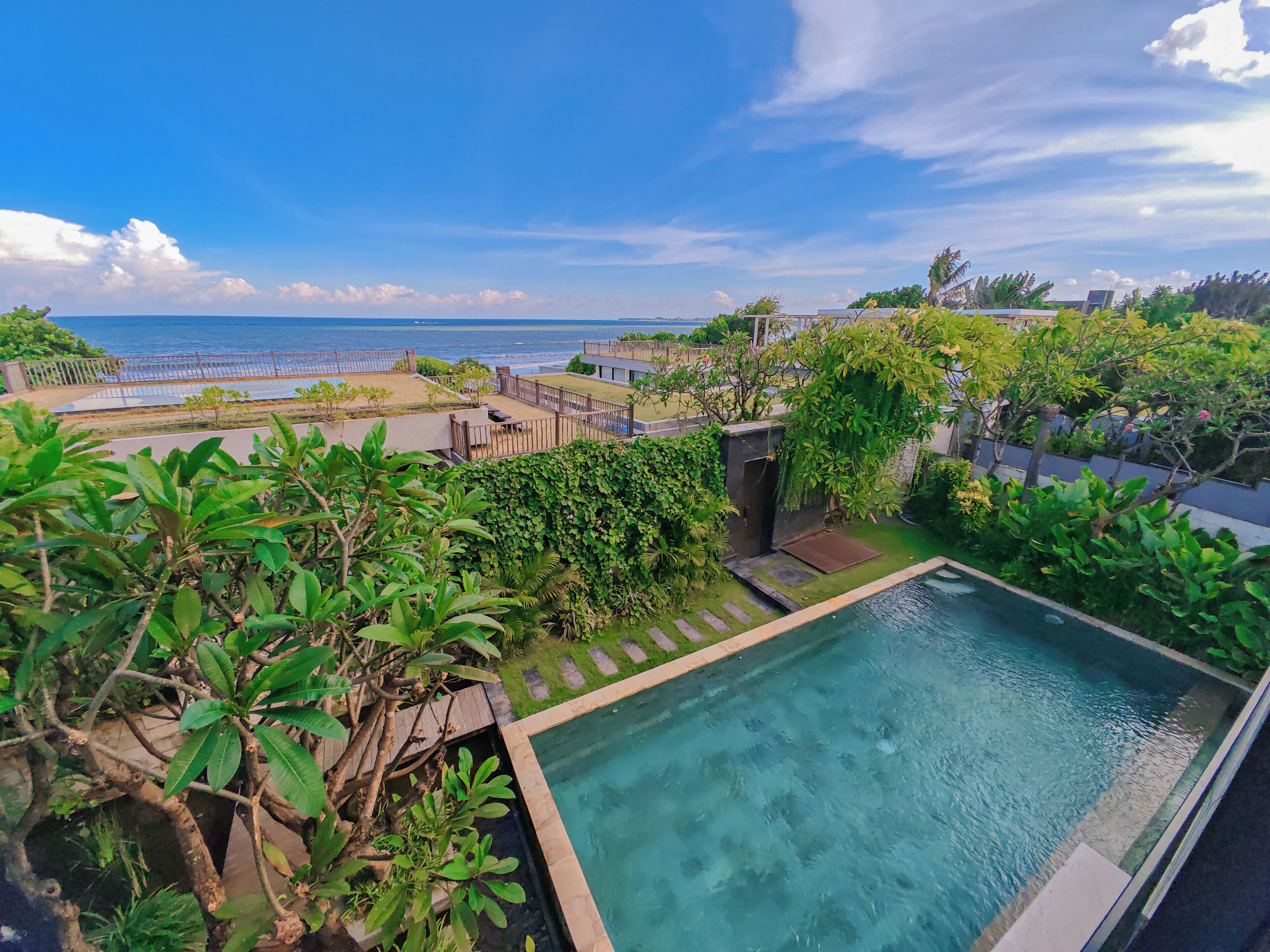 A rooftop pool with a view of the ocean, surrounded by greenery and a fence.