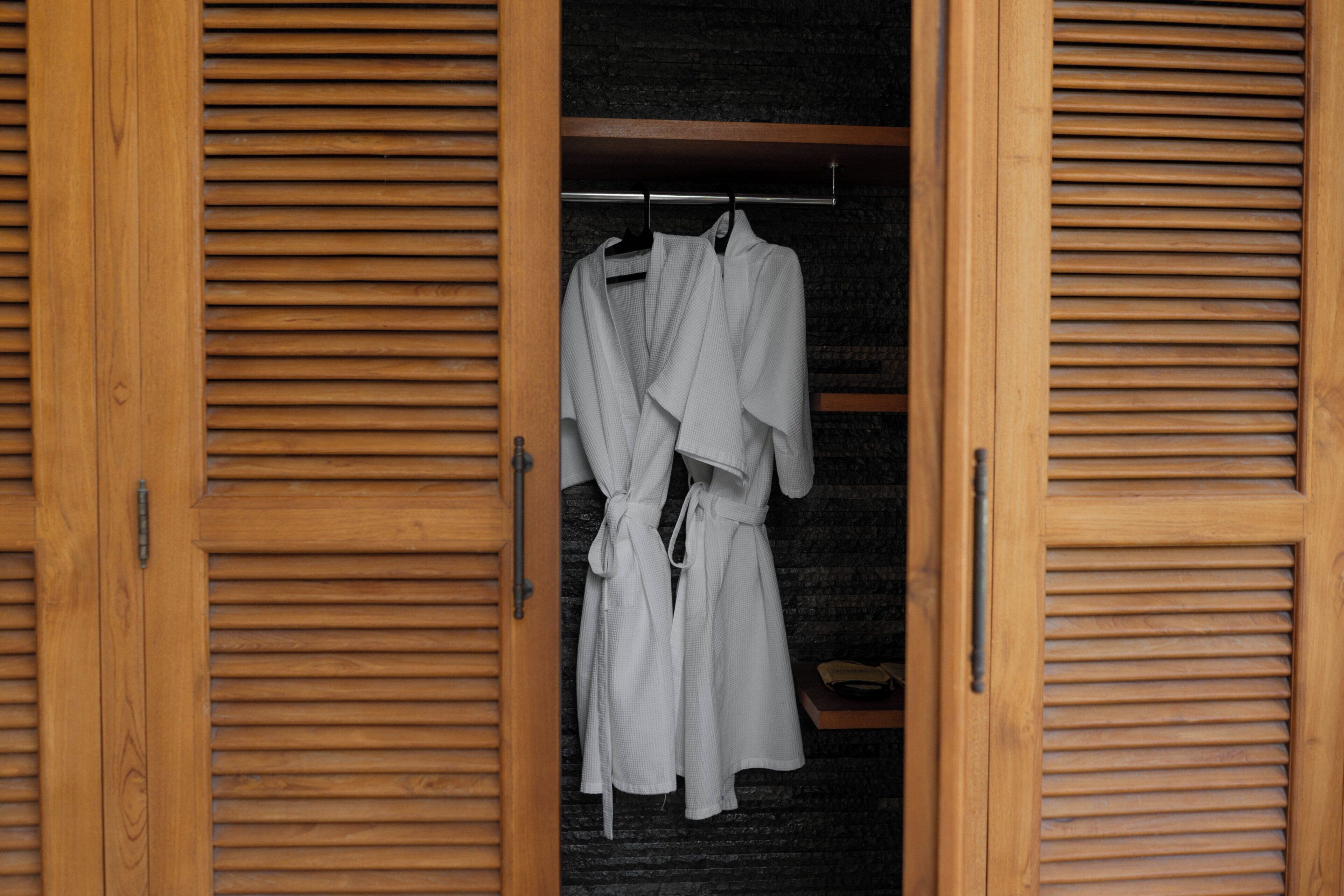 A wooden wardrobe with a hanging rack inside, containing a white robe and a white shirt.