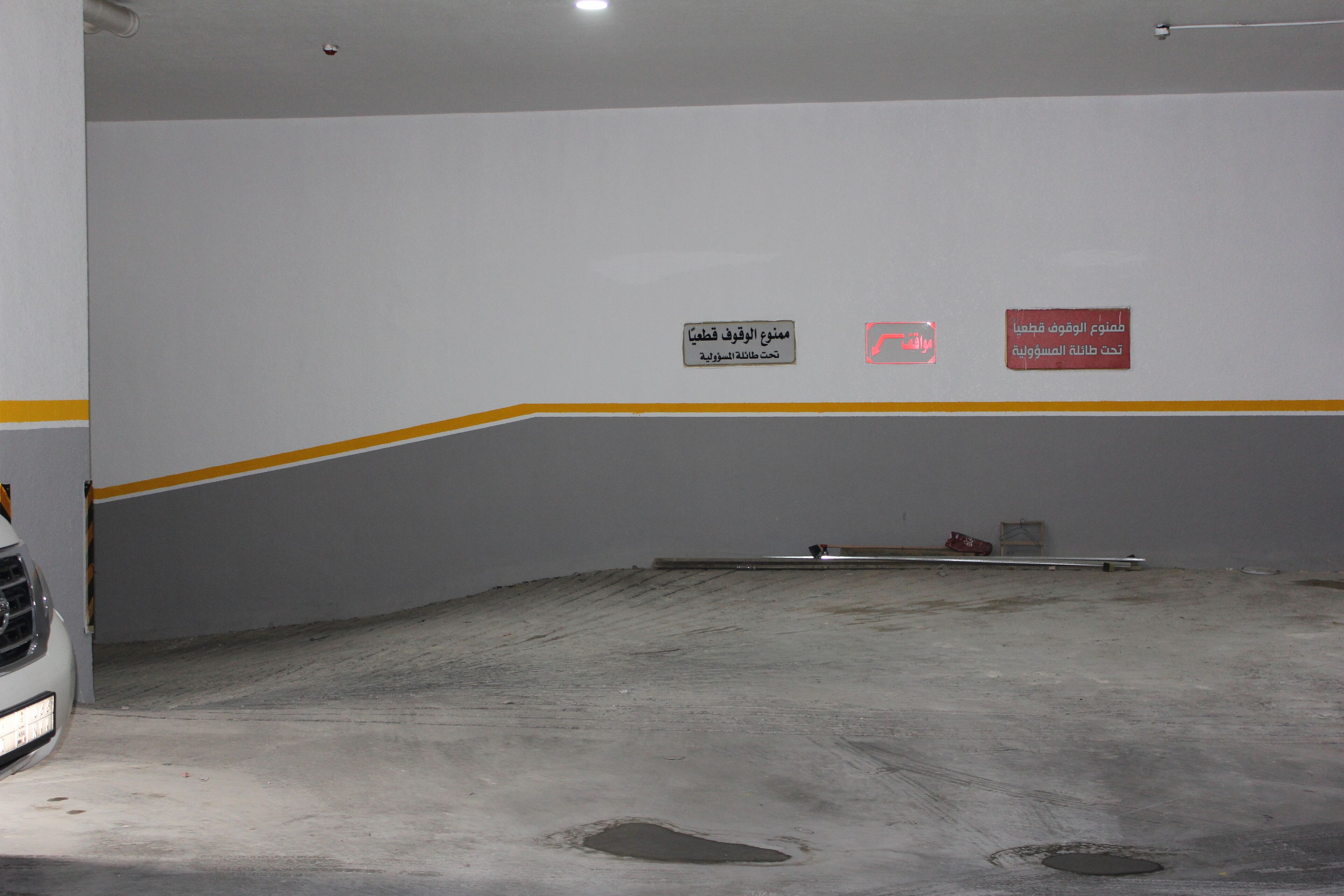 Un aparcamiento con pared blanca y franja amarilla, un coche blanco estacionado, y señales en árabe en la pared.