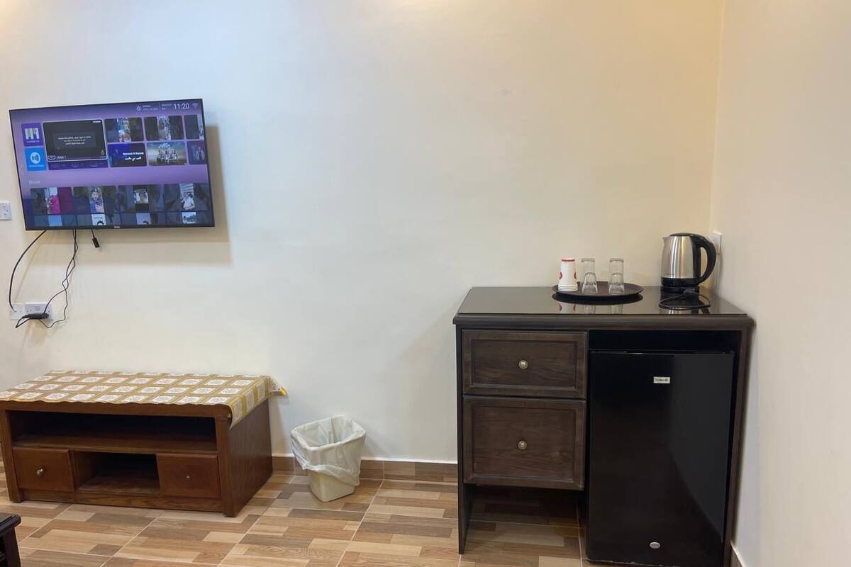 A room with a wooden bench, a small trash bin, a black refrigerator, and a television mounted on the wall displaying a video conference interface.