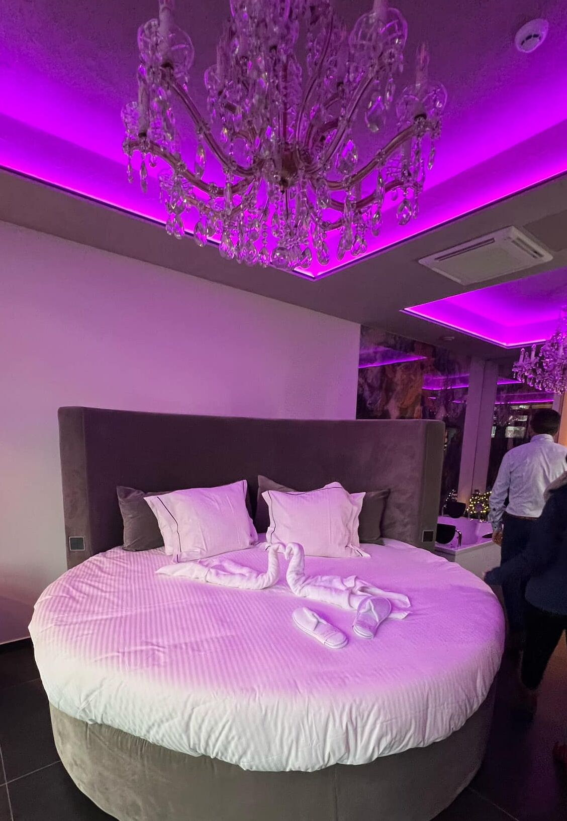 A large bed with a white and gray color scheme, a chandelier, and a person walking in the background.